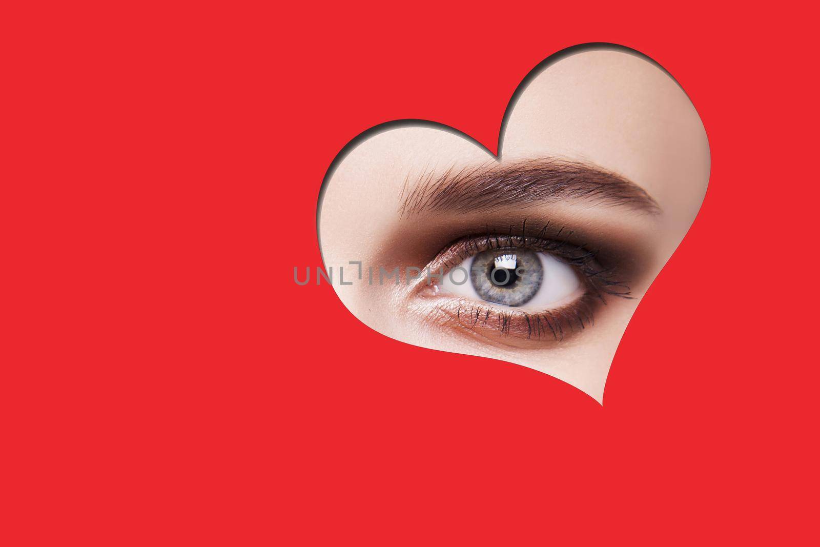 Young beautiful woman with smoky eyes makeup looking at camera through red heart shape. indoor studio shot isolated on red background.