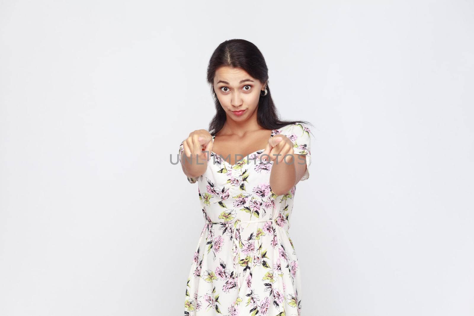 Hey you! Young adult woman, pointing finger and looking at camera. On gray background. Indoor, studio shot