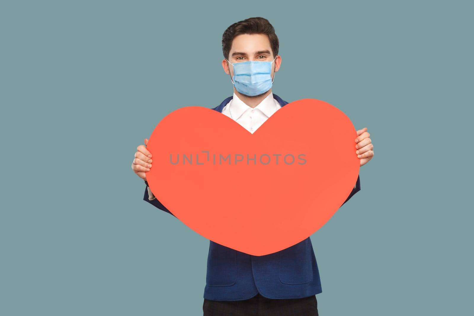happy young man with surgical medical mask standing, holding red big heart shape. looking at camera with good feeling. medicine and health care concept. Indoor, studio shot isolated on blue background