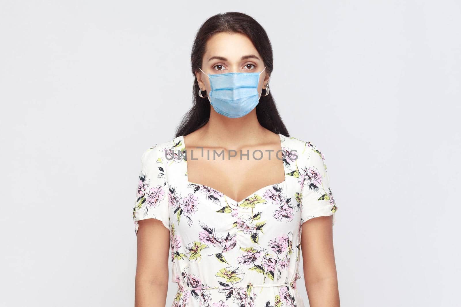 Protection against contagious disease, coronavirus. Portrait of calm young woman with surgical medical mask in white dress standing and looking at camera. indoor studio shot isolated, gray background.