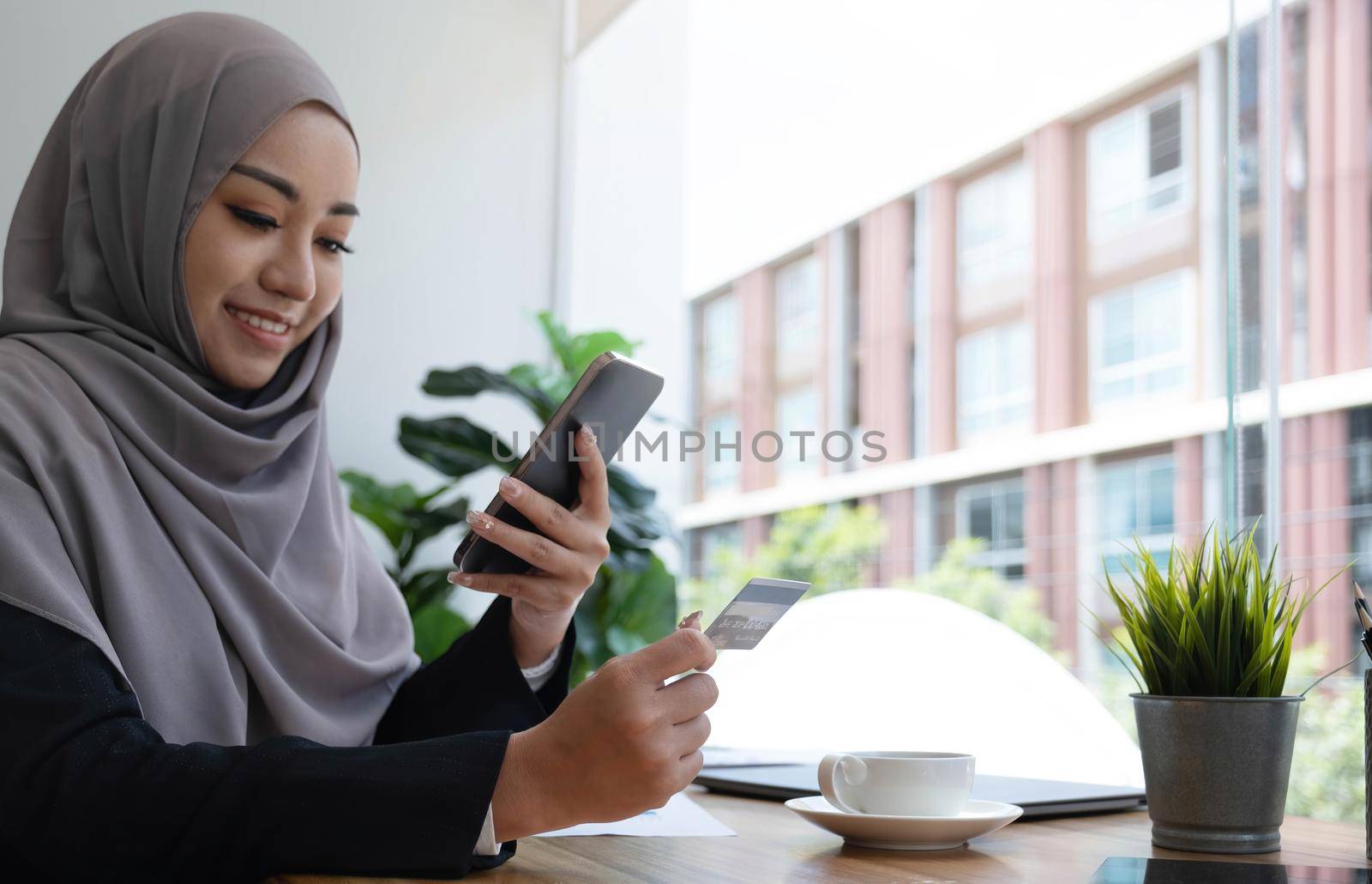 Beautiful young muslim woman with hijab sits at her office desk, holding a smartphone and credit card. Online payment, internet banking, online shopping concept. Close-up image.