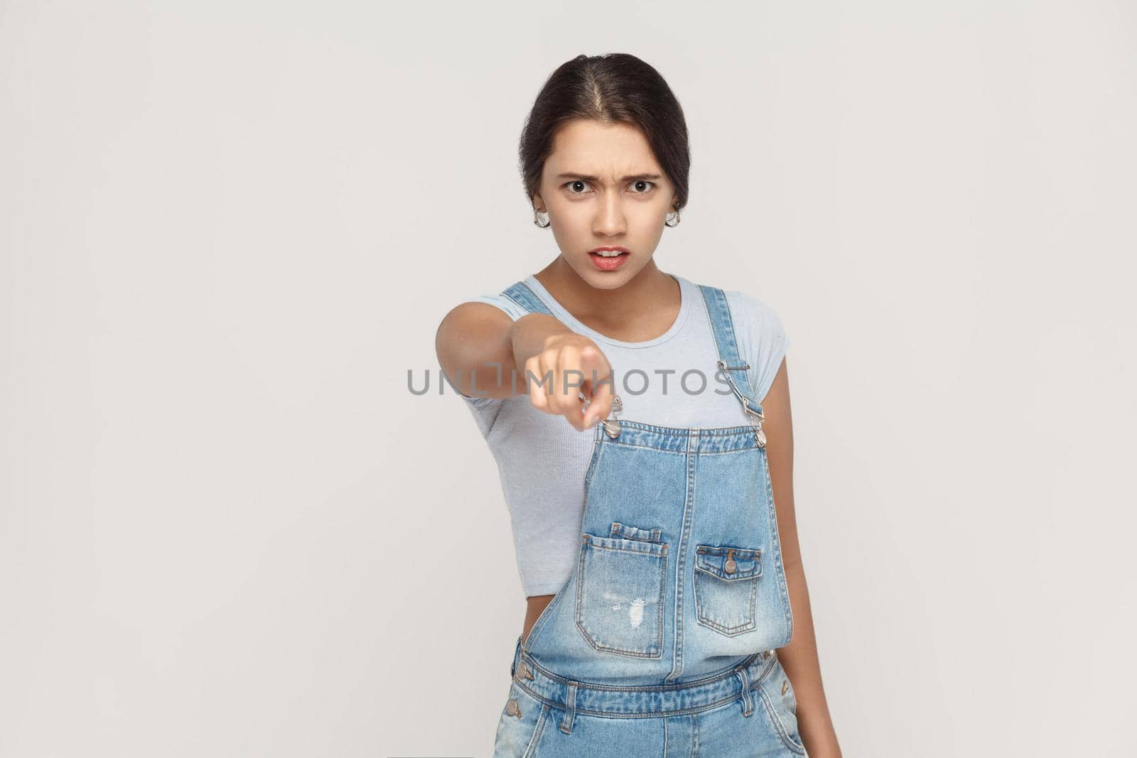 Angry latin woman with denim overalls looking at camera and pointing finger. Studio shot isolated on gray background.
