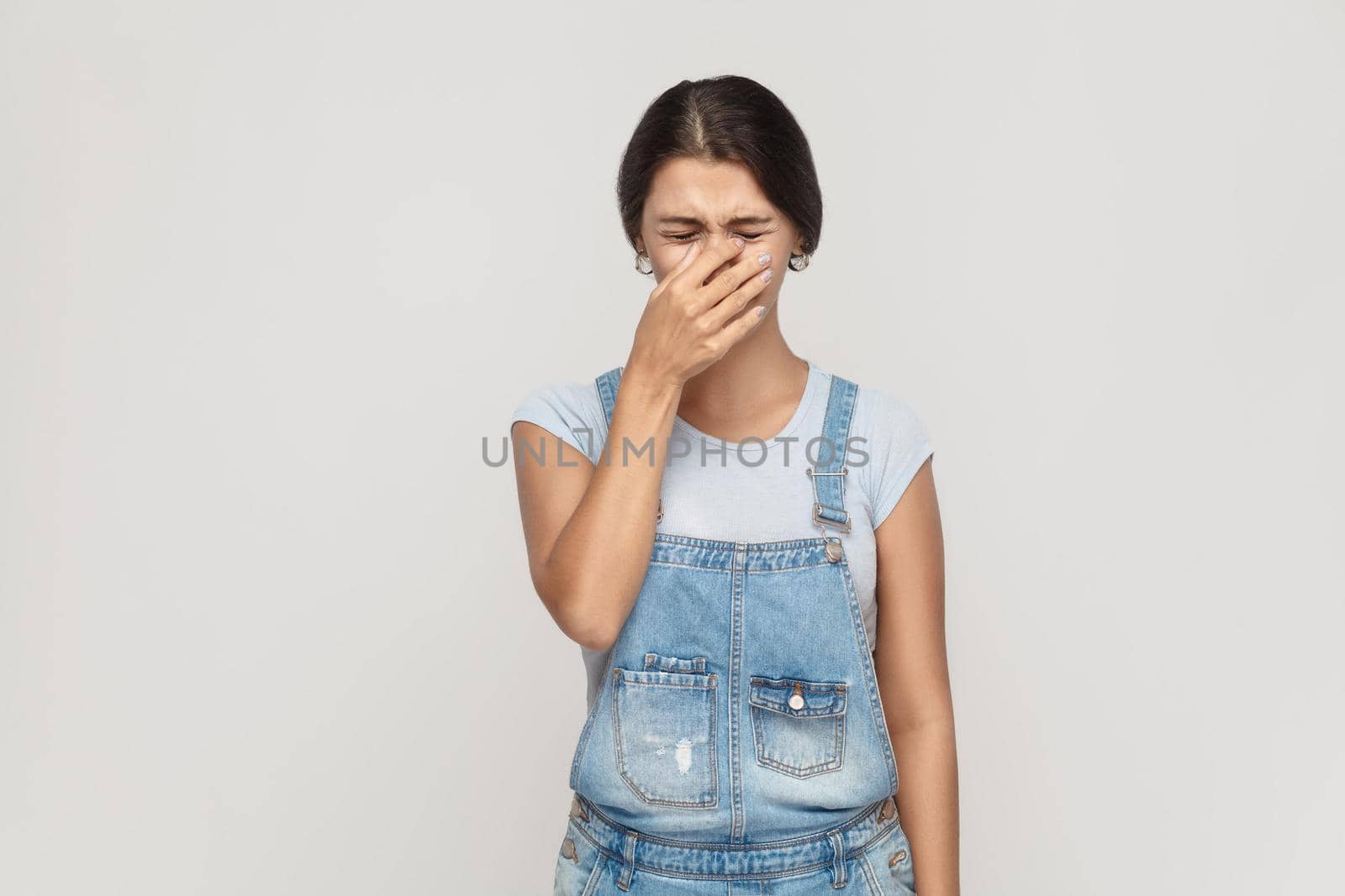 Unhappy and depressed young adult gypsy woman, feeling ashamed or sick, covering face with both hands, keeping eyes closed. Isolated studio shot on gray background.
