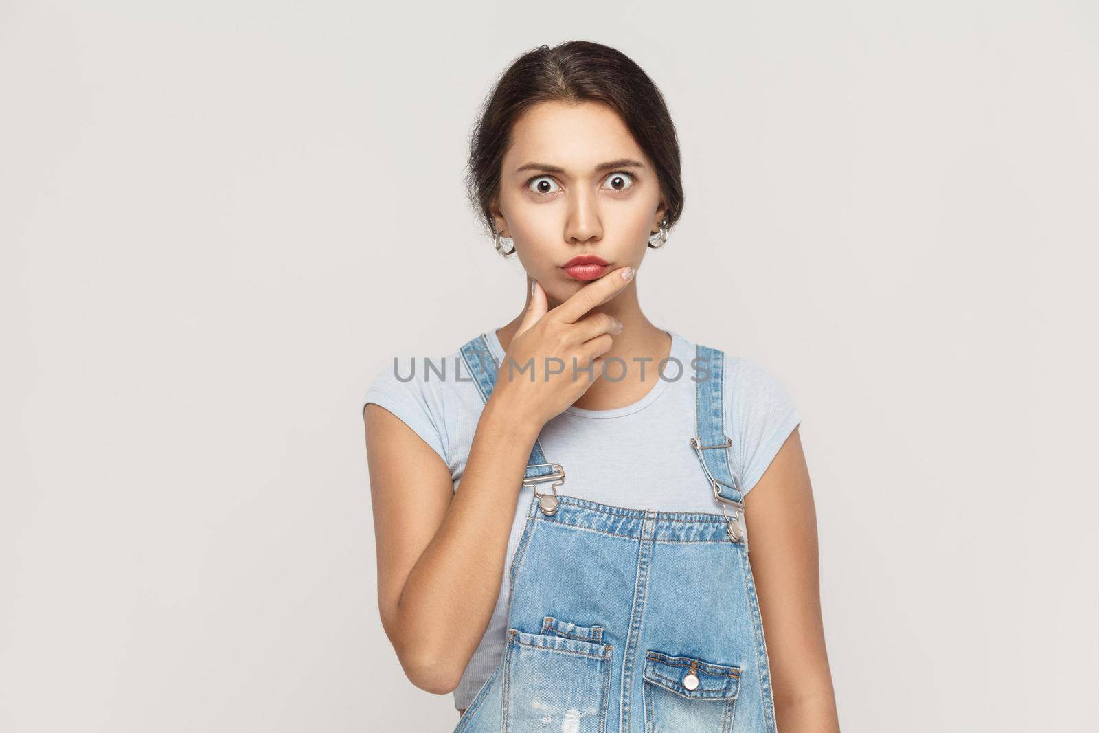 Amazed hispanic woman looking at camera with big eyes and touching her chin. Studio shot, isolated on gray background
