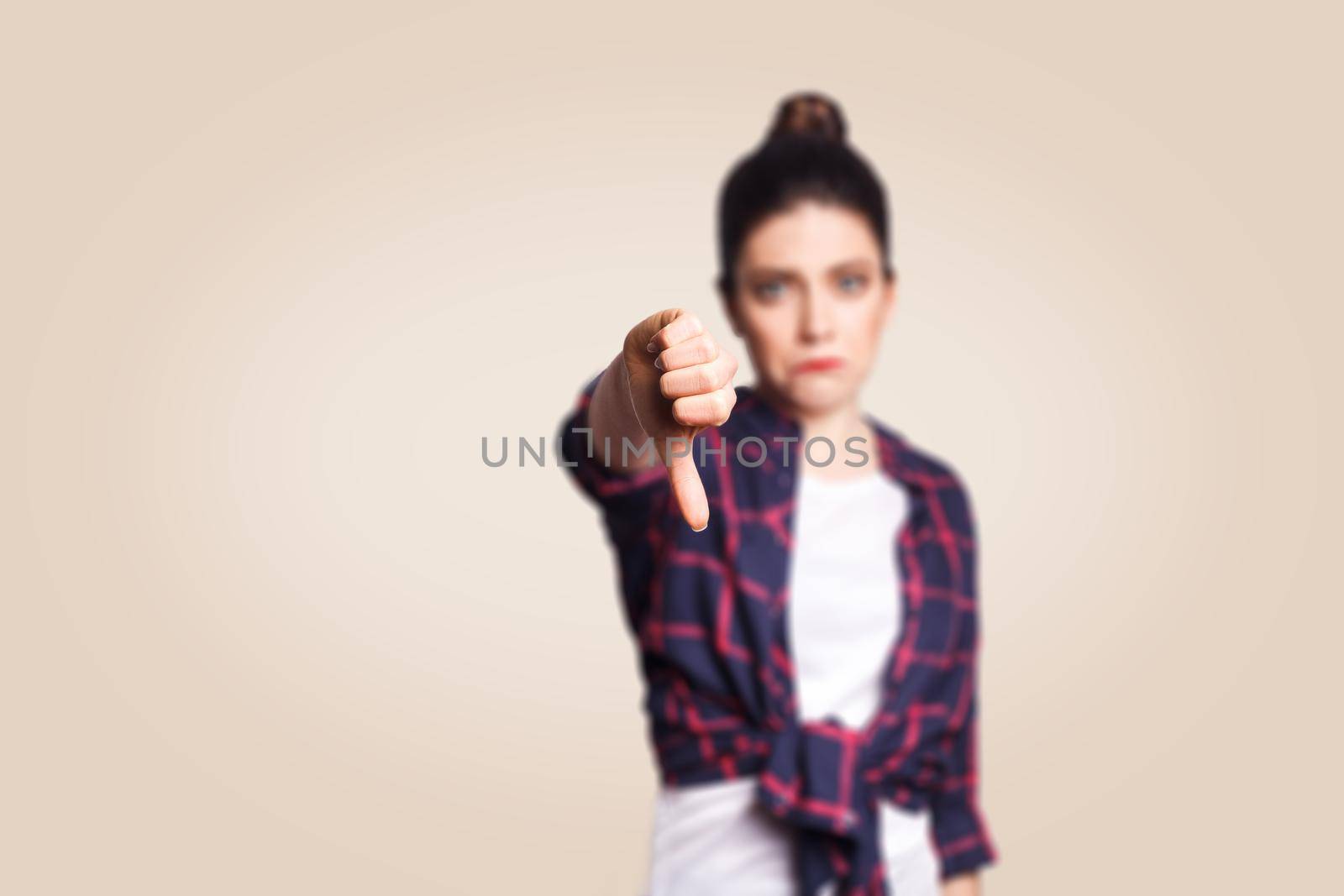 Dislike. Young unhappy upset girl with casual style and bun hair thumbs down her finger, on beige blank wall with copy space looking at camera with toothy smile. focus on finger.