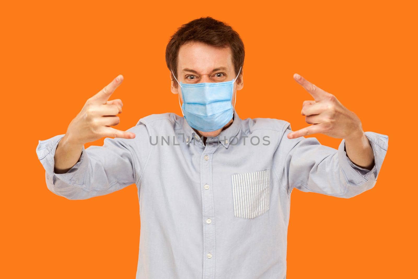 Yes. Portrait of young worker man with surgical medical mask standing with rock horns and loolong at camera screaming. indoor studio shot isolated on orange background.