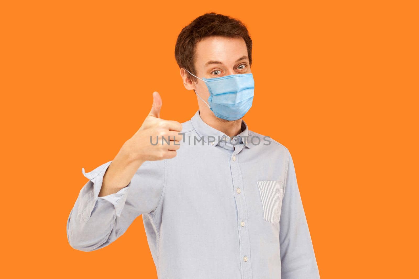 Like. Portrait of satisfied young worker man with surgical medical mask standing thumbs up and looking at camera smiling. indoor studio shot isolated on orange background.
