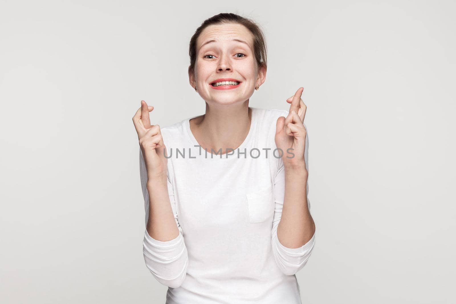 Young adult beautiful girl toothy smiling and looking at camera, crossed fingers. Studio shot,isolated on gray background