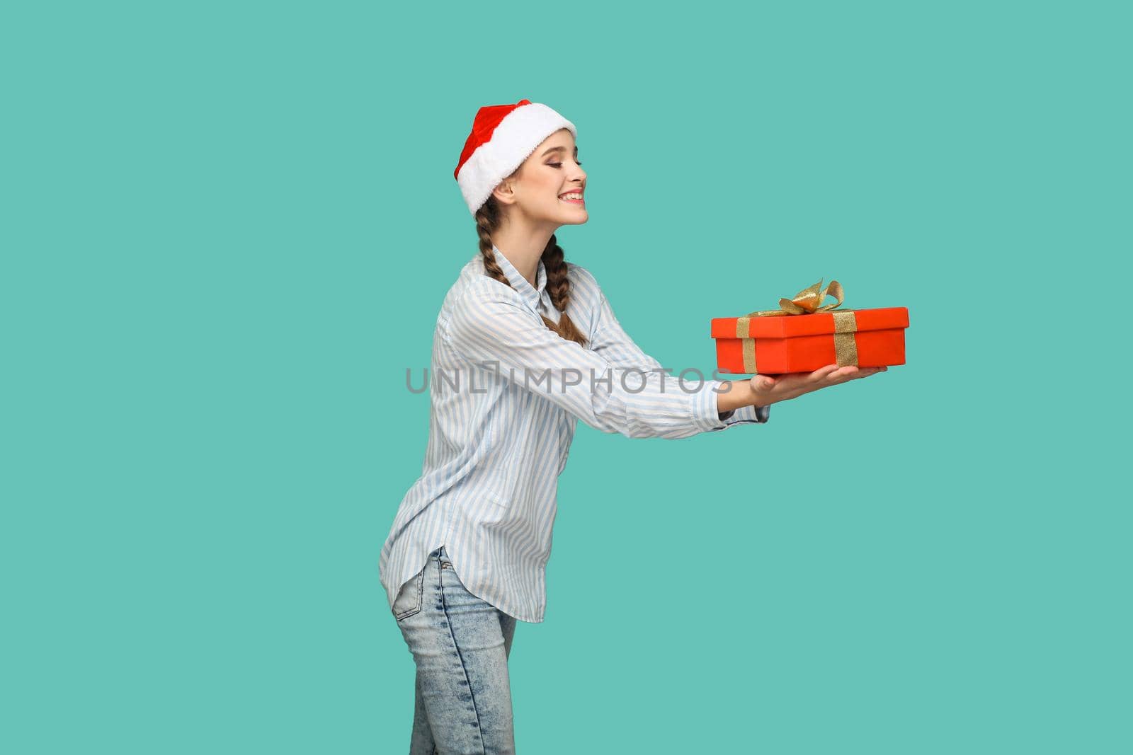 New year concept. profile side view of beautiful girl in striped light blue shirt in red christmas cap standing and giving red gift box with toothy smile. indoor, isolated on green background.