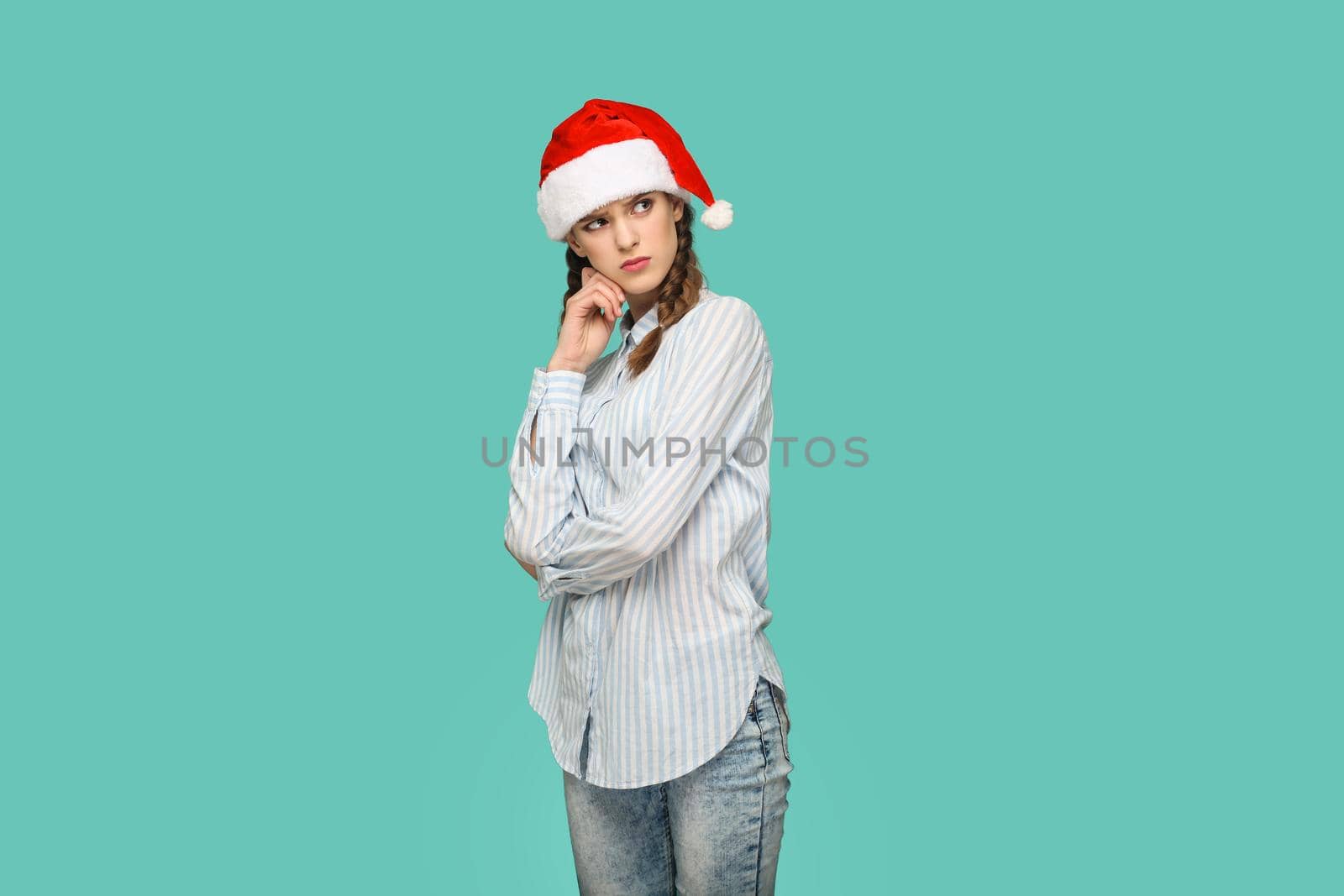 New year concept. thoughtful beautiful girl in striped light blue shirt in red christmas cap standing crossed hands, confused face and thinking. indoor, studio shot isolated on green background.