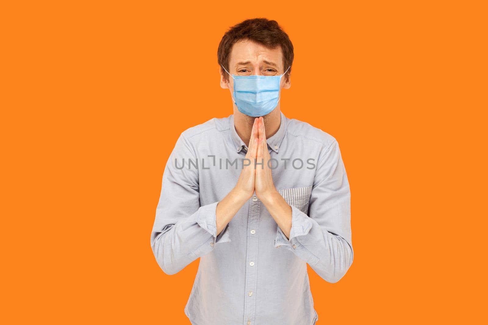 Please help me. Portrait of sad worry young worker man with medical mask standing with palm hands, worry and looking at camera begging or pleading. indoor studio shot isolated on orange background.