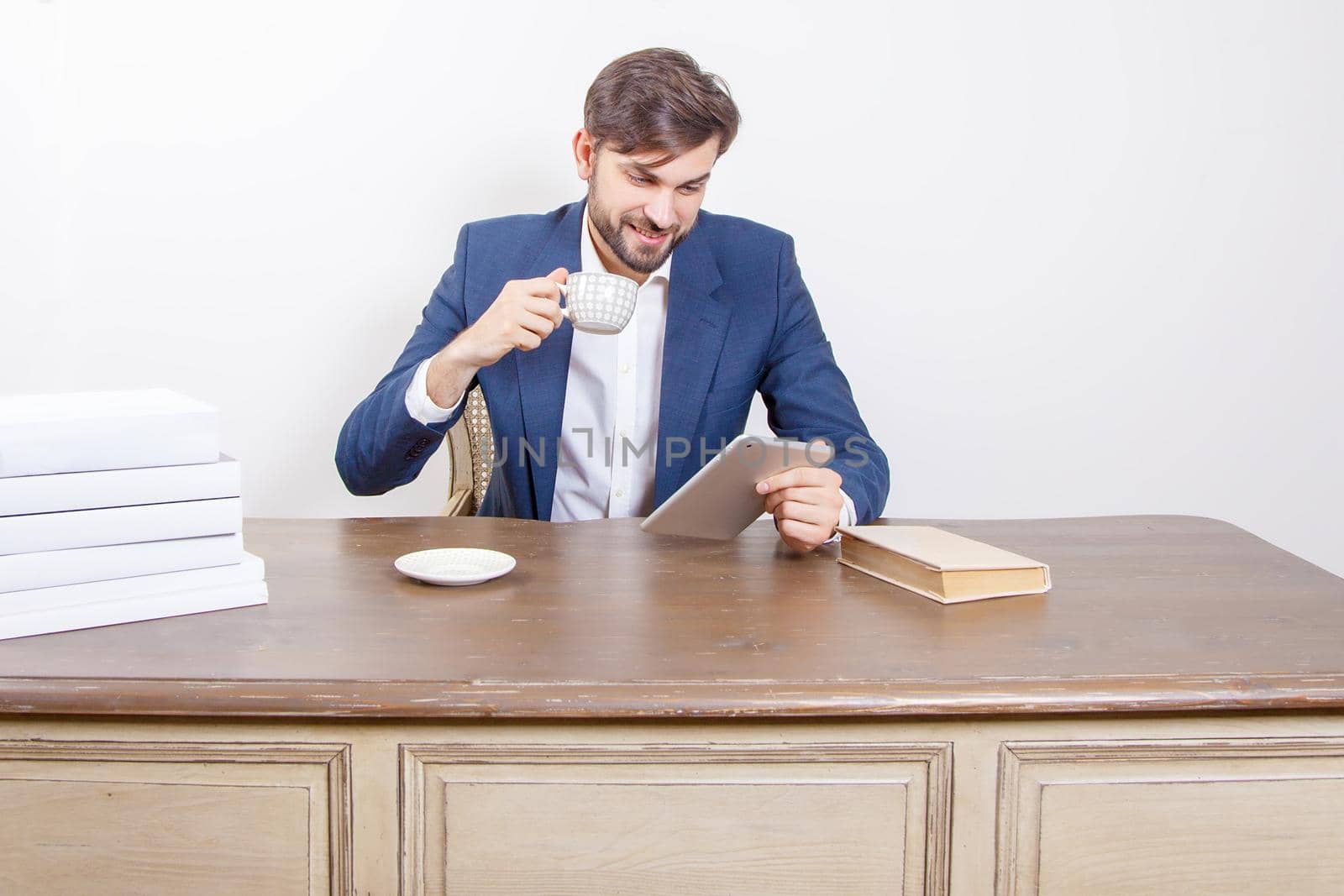 Technology, people and business concept - handsome man with beard and brown hair and blue suit and tablet pc computer and some books and cup drinking coffee or tea sitting in the office holding tablet and smiling. .Isolated on white background. .