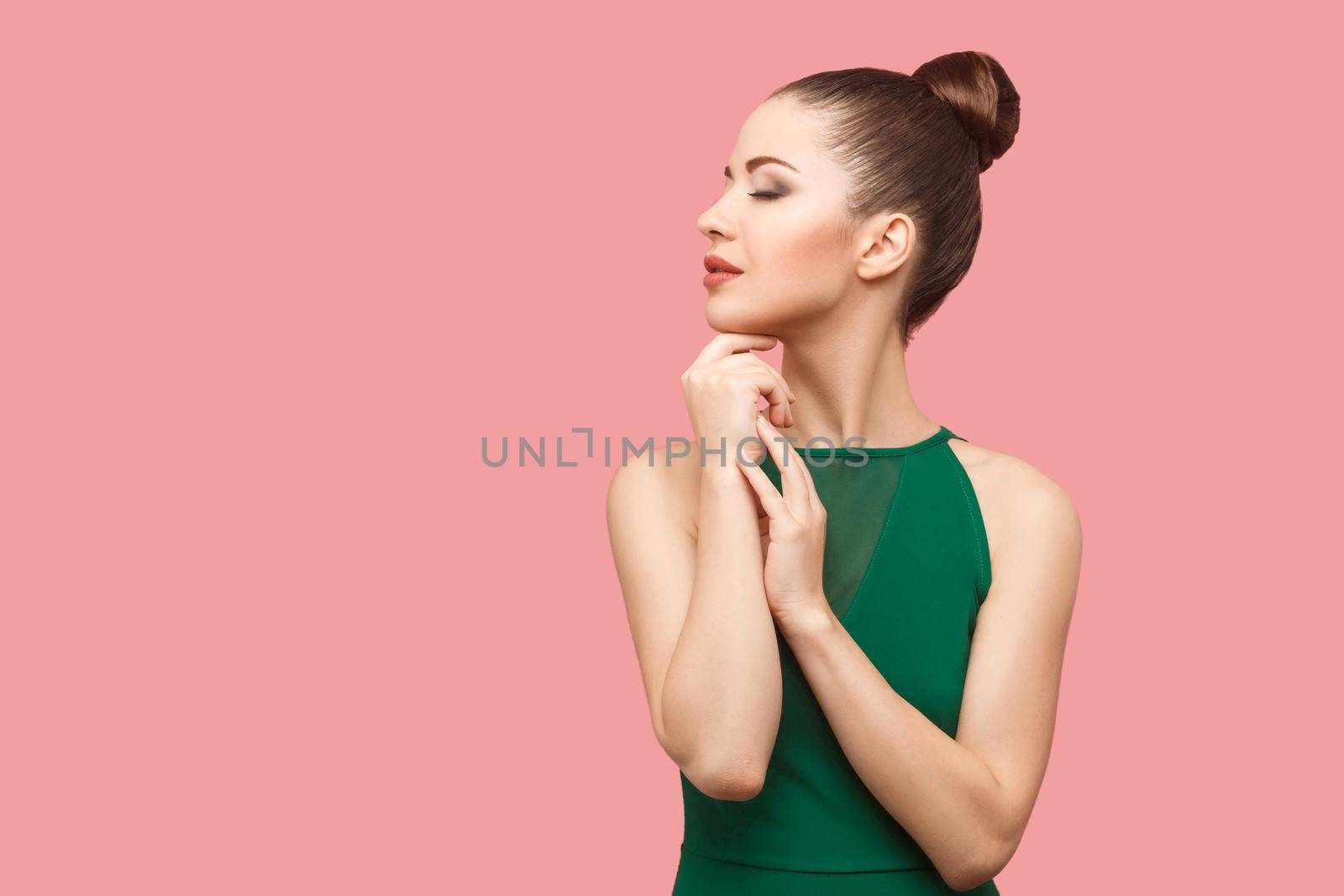 Profile side view portrait of calm serious beautiful young woman with bun hairstyle and makeup in green dress standing with closed eyes and touching her chin. studio shot, isolated on pink background.