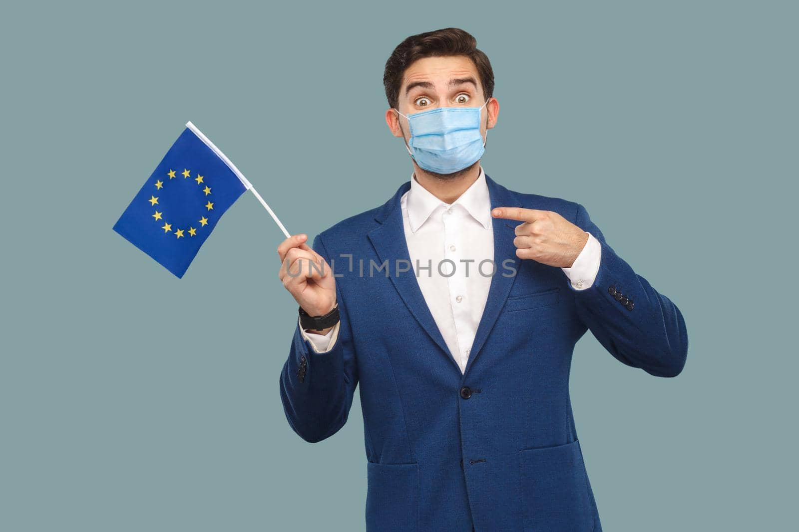 Young man with surgical medical mask in blue jacket holding and pointing at European union flag and looking at camera with big eyes and shocked faee. Indoor, studio shot isolated on blue background.