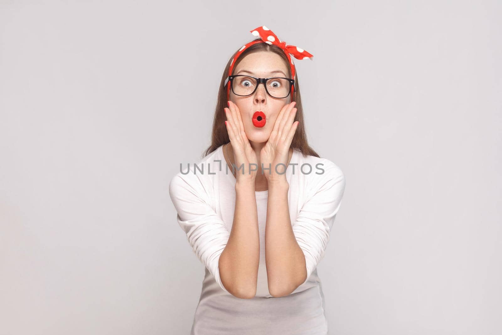 surprised big eyes portrait of beautiful emotional young woman in white t-shirt with freckles, black glasses, red lips and head band. indoor studio shot, isolated on light gray background.