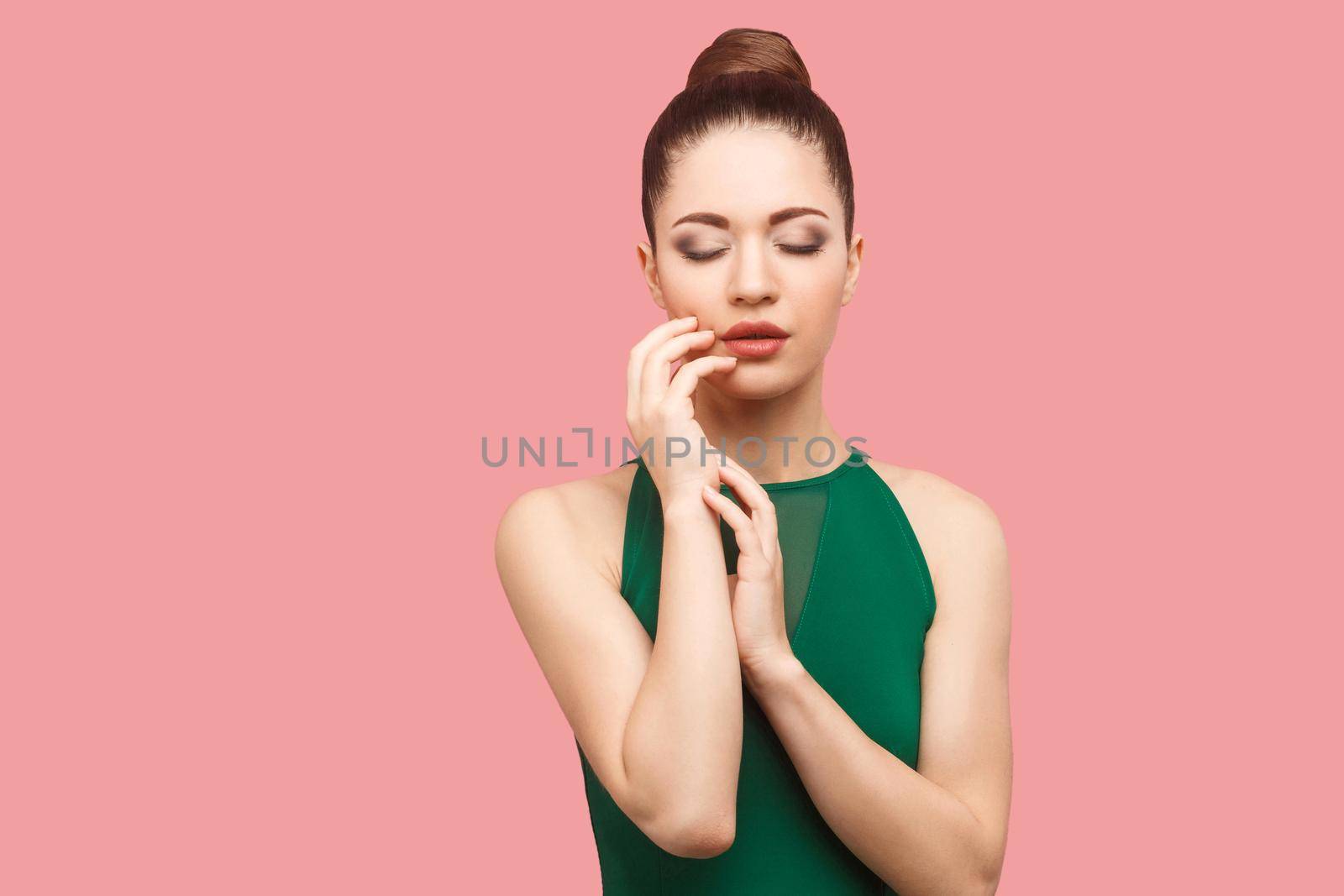 Beauty portrait of calm serious beautiful young woman with bun hairstyle and makeup in green dress standing, with closed her eyes and touching her face. indoor studio shot, isolated on pink background