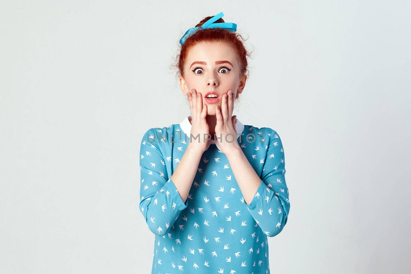 Human face expressions and emotions. Redhead young girl touching her cheeks and looking at camera with shocked face. Isolated studio shot on gray background.