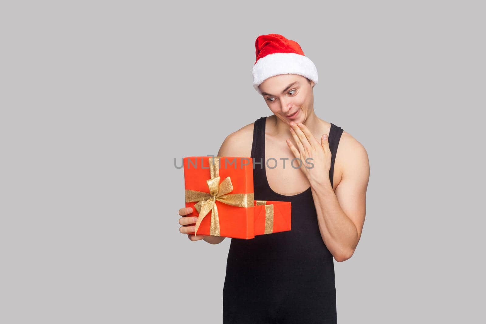 New year concept. Man in red hat holding red gift box and looking inside with amazed funny face. Indoor, studio shot, isolated on gray background