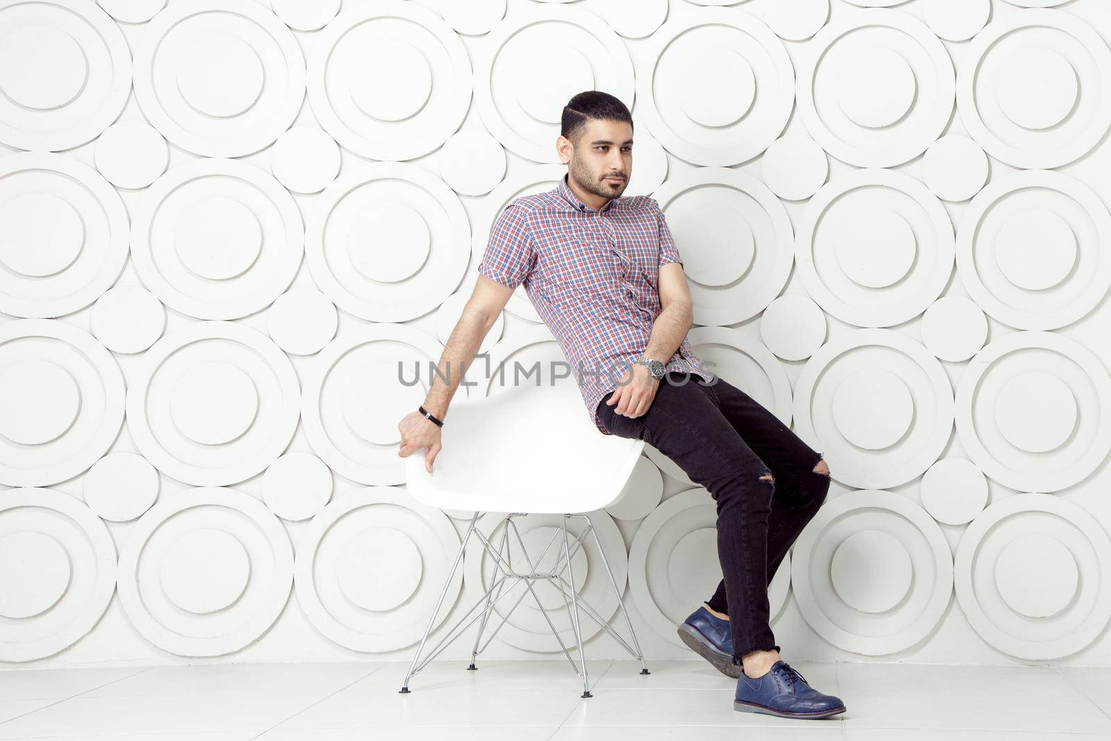 Young bearded fashion model in casual style is posing near white circle wall background. studio shot. sitting on chair and looking at away.
