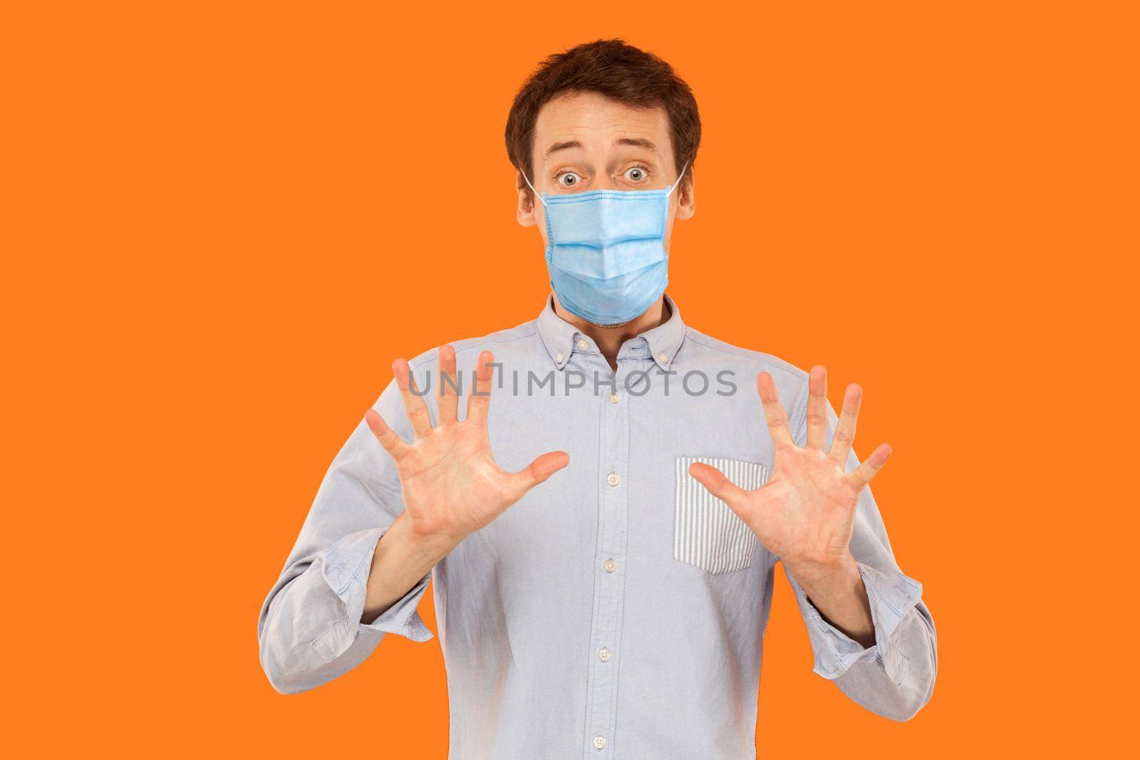 Portrait of scared young worker man with surgical medical mask standing blocking with hands and looking at camera with shocked afraid face. indoor studio shot isolated on orange background.