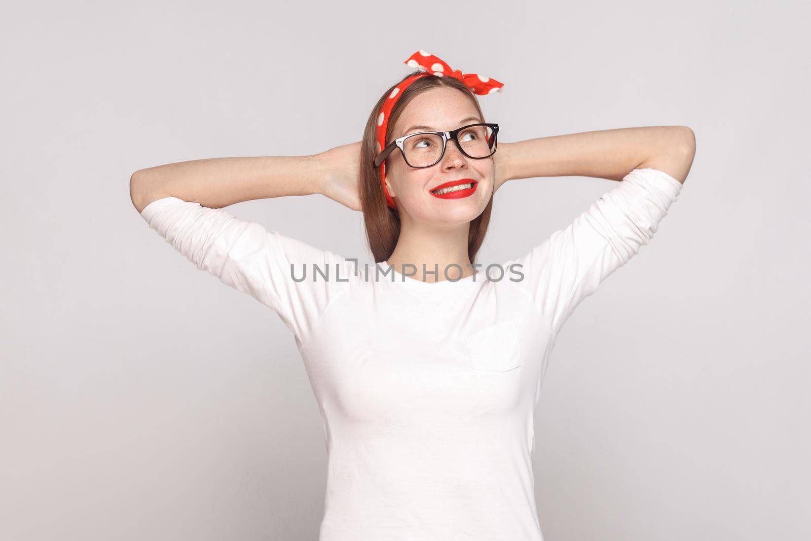 dreaming hopeful portrait of beautiful emotional young woman in white t-shirt with freckles, black glasses, red lips and head band. indoor studio shot, isolated on light gray background.