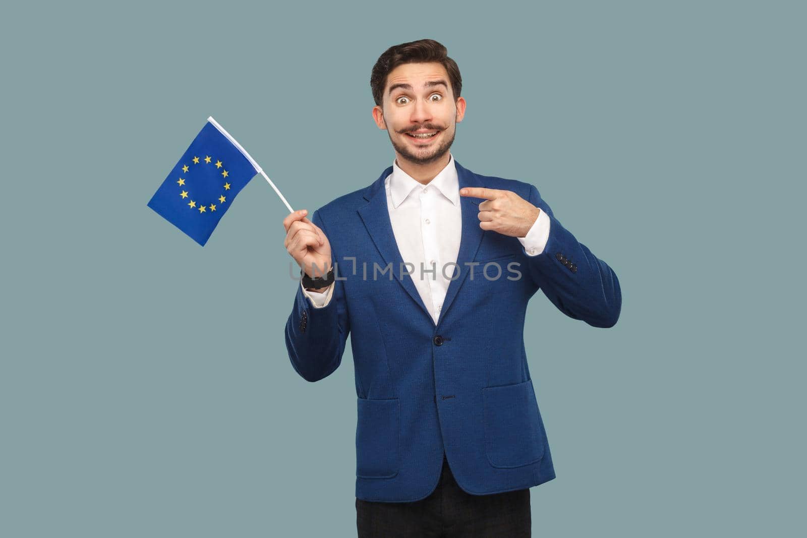 Handsome businessman in blue jacket and white shirt holding and pointing finger at European union flag and looking at camera with toothy smile. Indoor, studio shot isolated on light blue background.