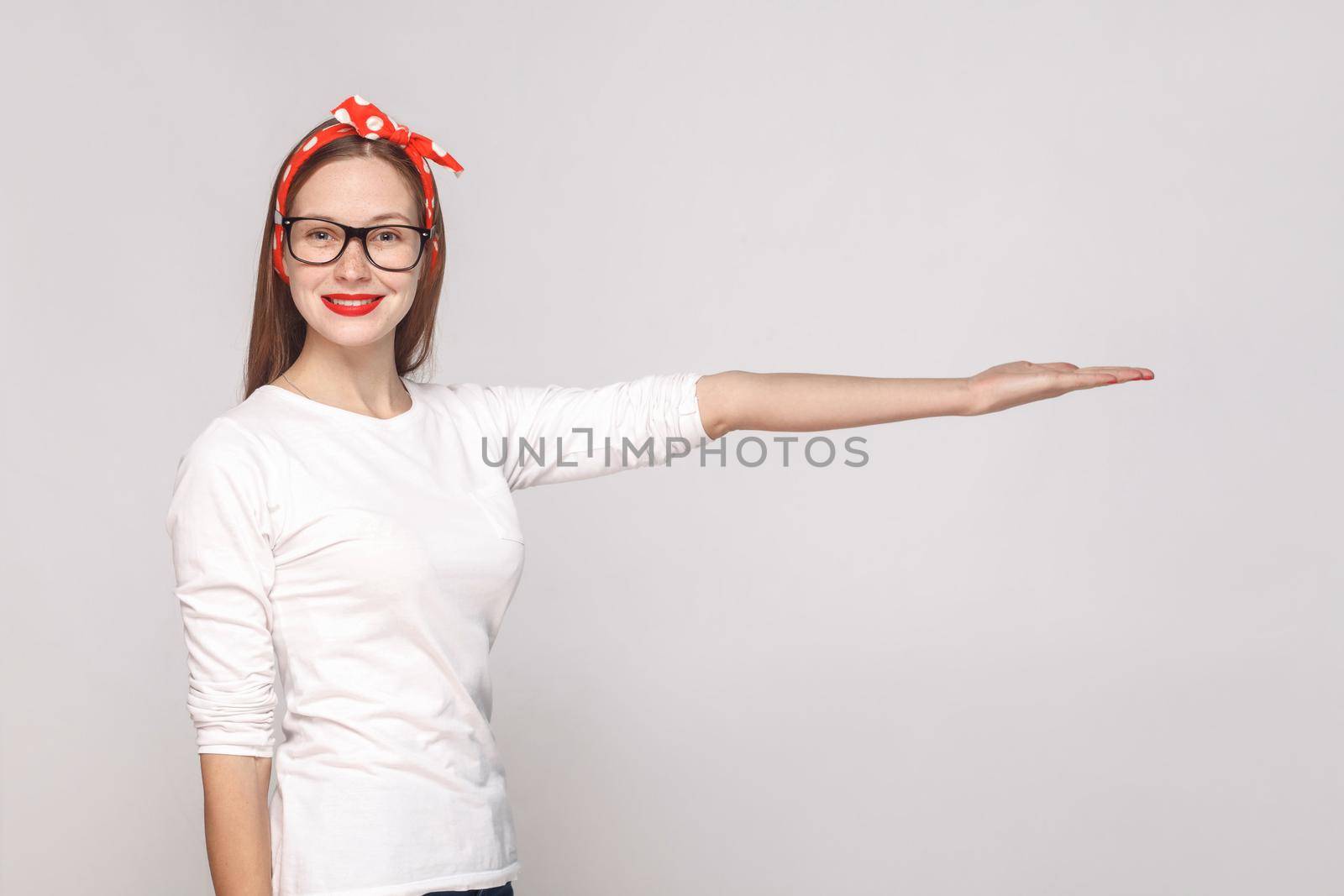 portrait of beautiful emotional young woman in white t-shirt with glasses, red lips and head band looking at camera with toothy smile and showing copy space. indoor shot, isolated on gray background.