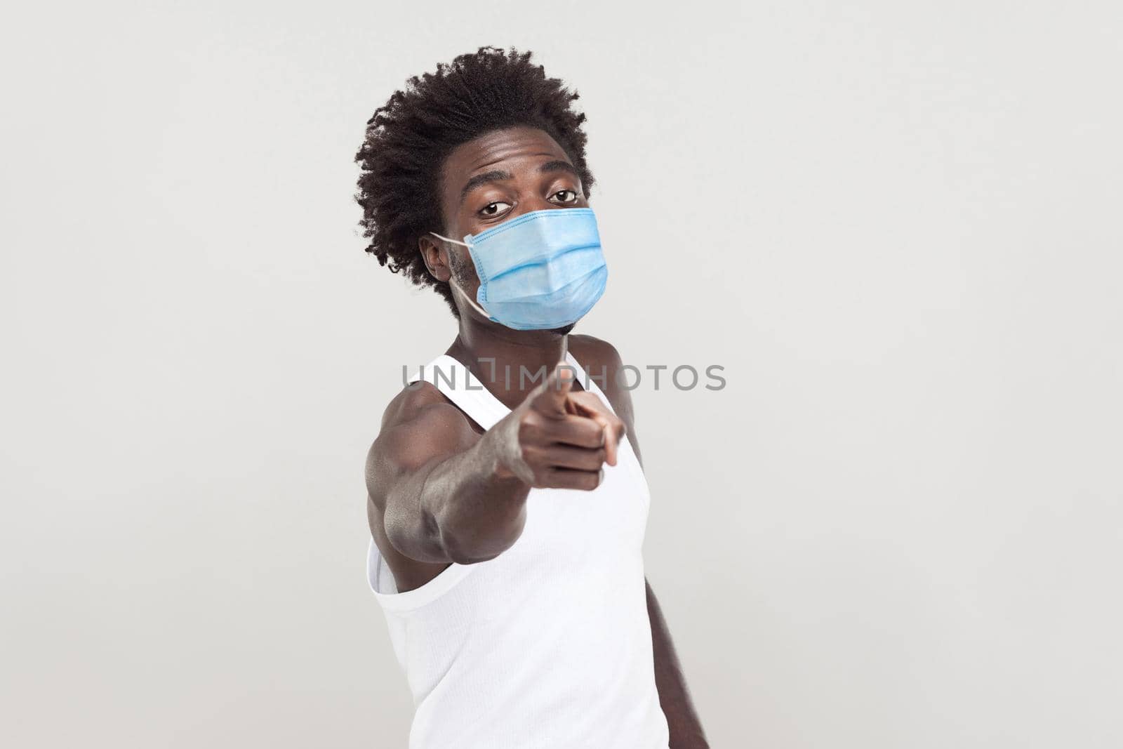 Hey you. Portrait of serious young man wearing white shirt with surgical medical mask standing looking and pointing at camera with serious face. indoor studio shot isolated on gray background.
