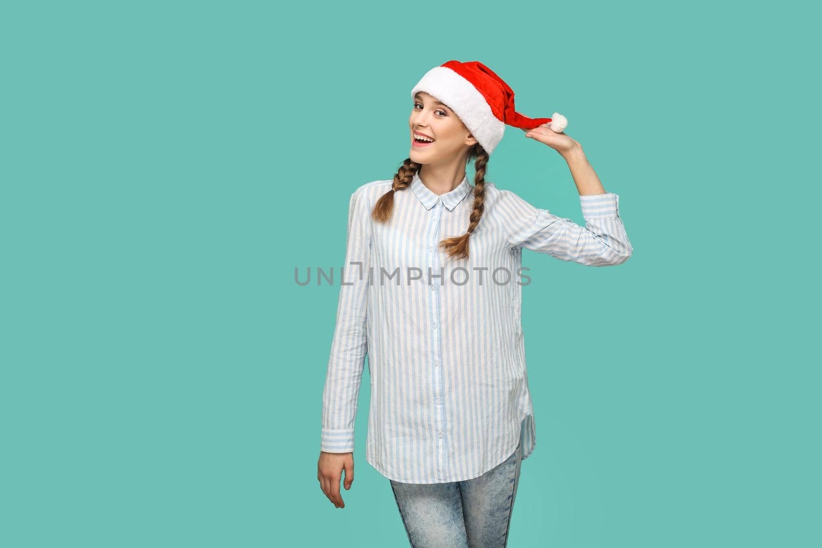 New year concept. happy funny beautiful girl in striped light blue shirt standing and holding red christmas hat, and looking at camera with toothy smile. indoor isolated on green background.