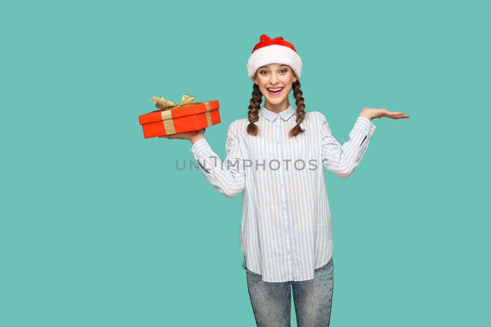 New year concept. happy beautiful girl in striped light blue shirt and red christmas cap holding red gift box and looking at camera with surprised face. indoor isolated on green background.
