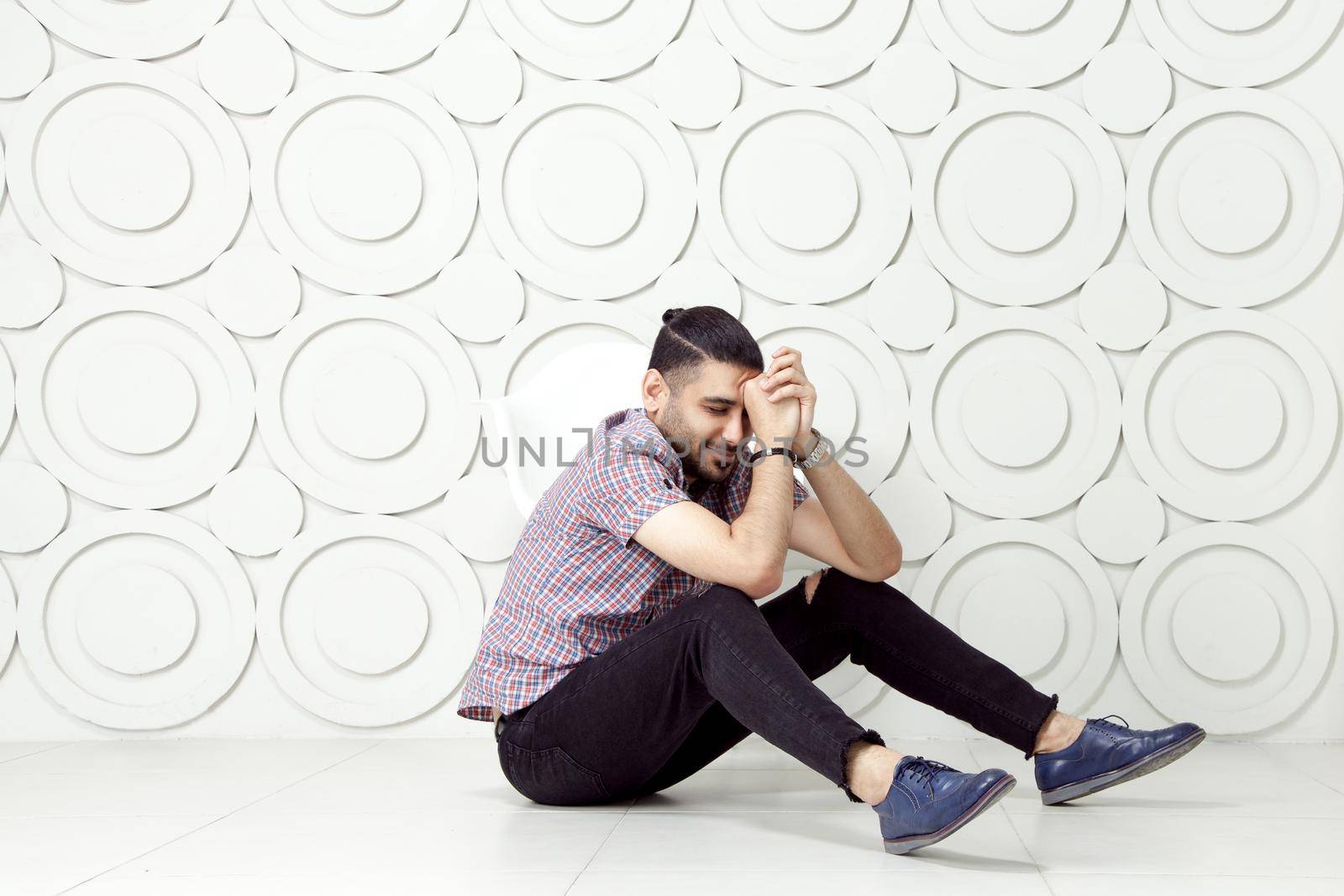 Young bearded fashion model in casual style is posing near white circle wall background. studio shot. sitting on floor and smiling with closed eyes.