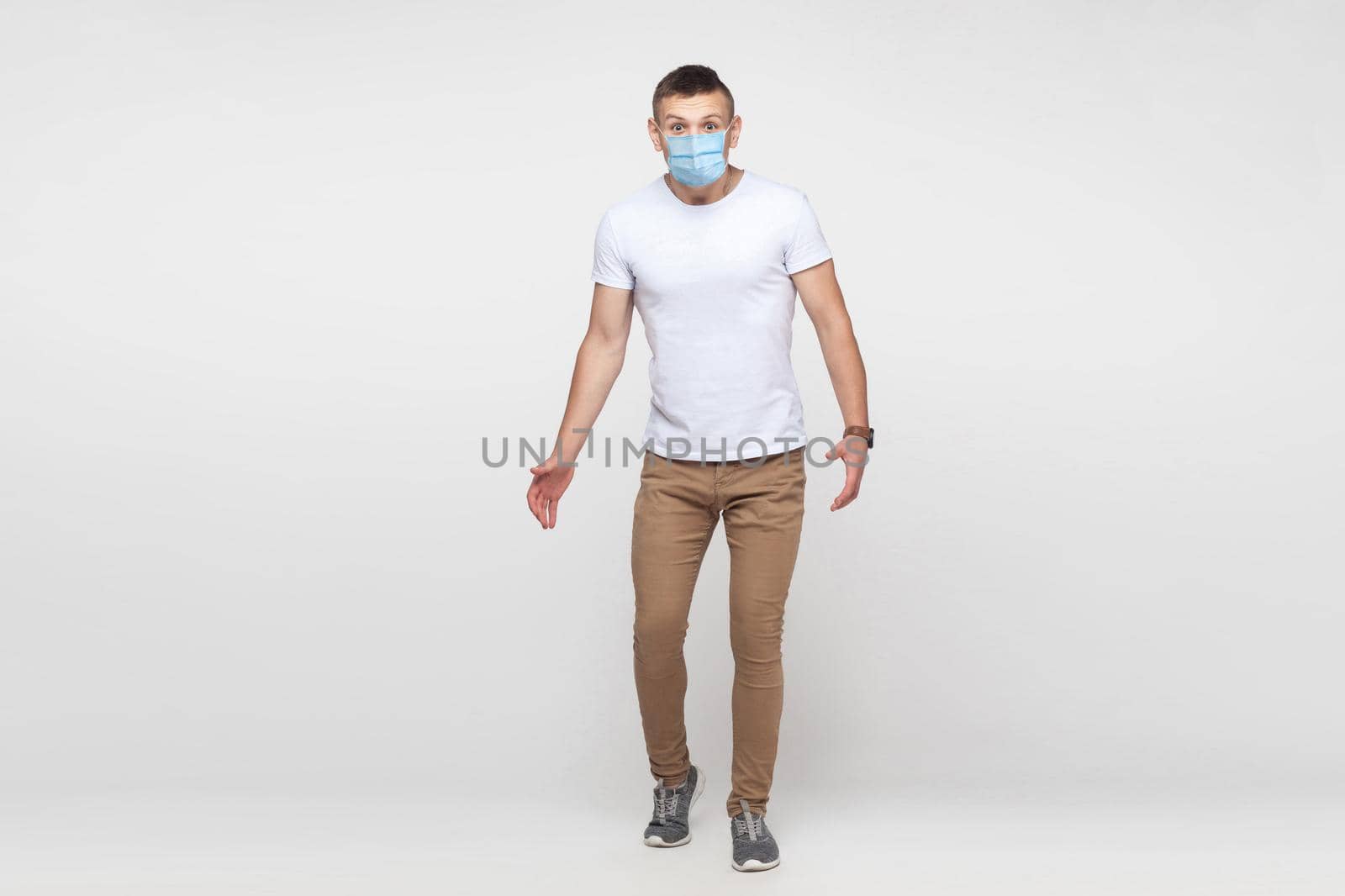 Full length portrait of amazed young man in white shirt with surgical medical mask standing with big eyes and looking at camera. indoor studio shot, isolated on gray background.