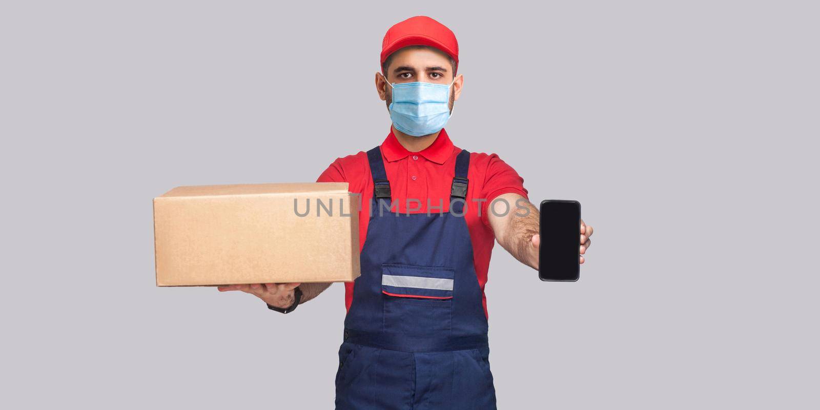 Delivery on quarantine. This is For you! Young man with surgical medical mask in blue uniform and red t-shirt standing, holding cardboard box and showing smart phone display on grey background.