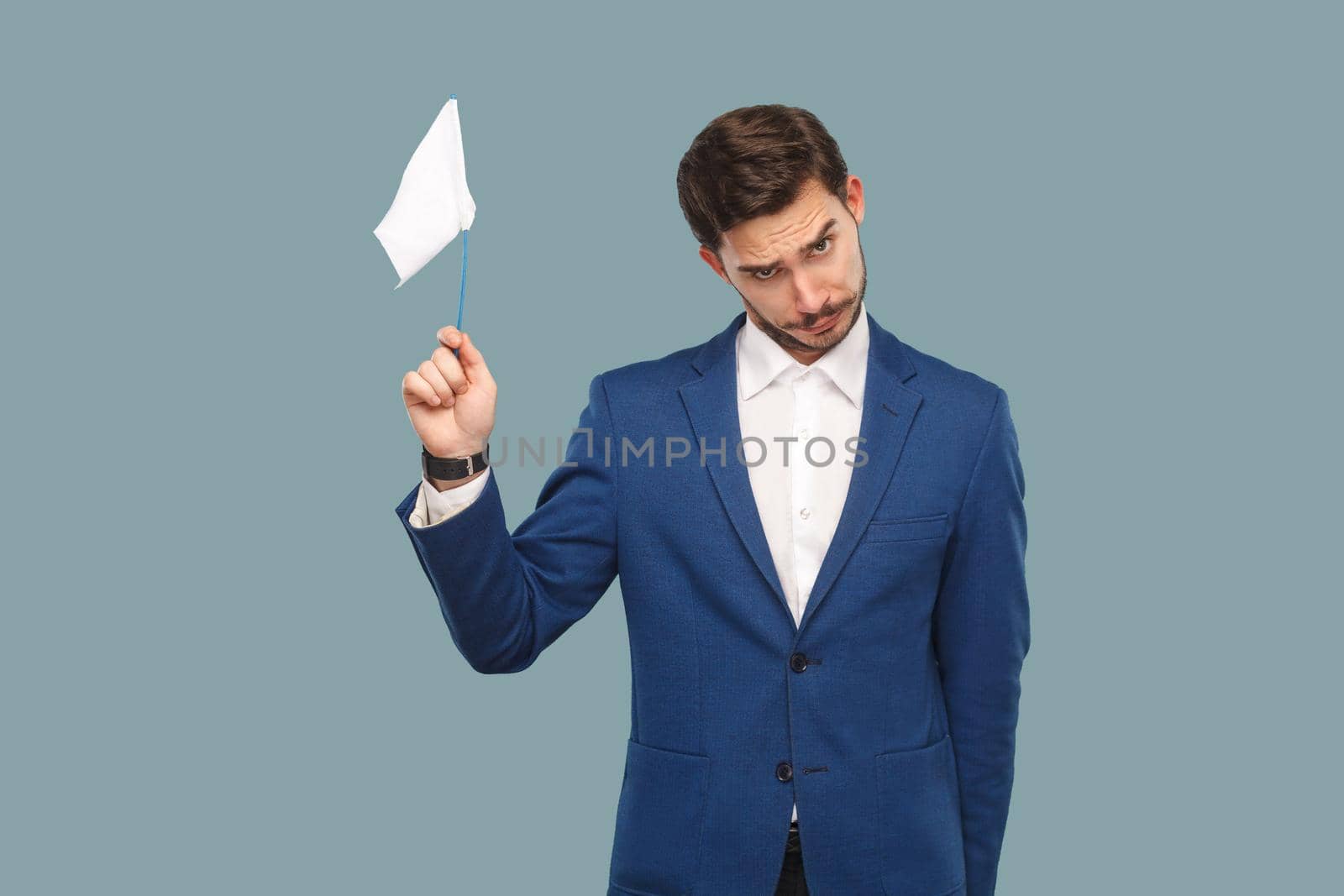 I am give up. sad failure businessman in blue jacket and white shirt standing and holding white flag and looking at camera with sad face. Indoor, studio shot isolated on light blue background.