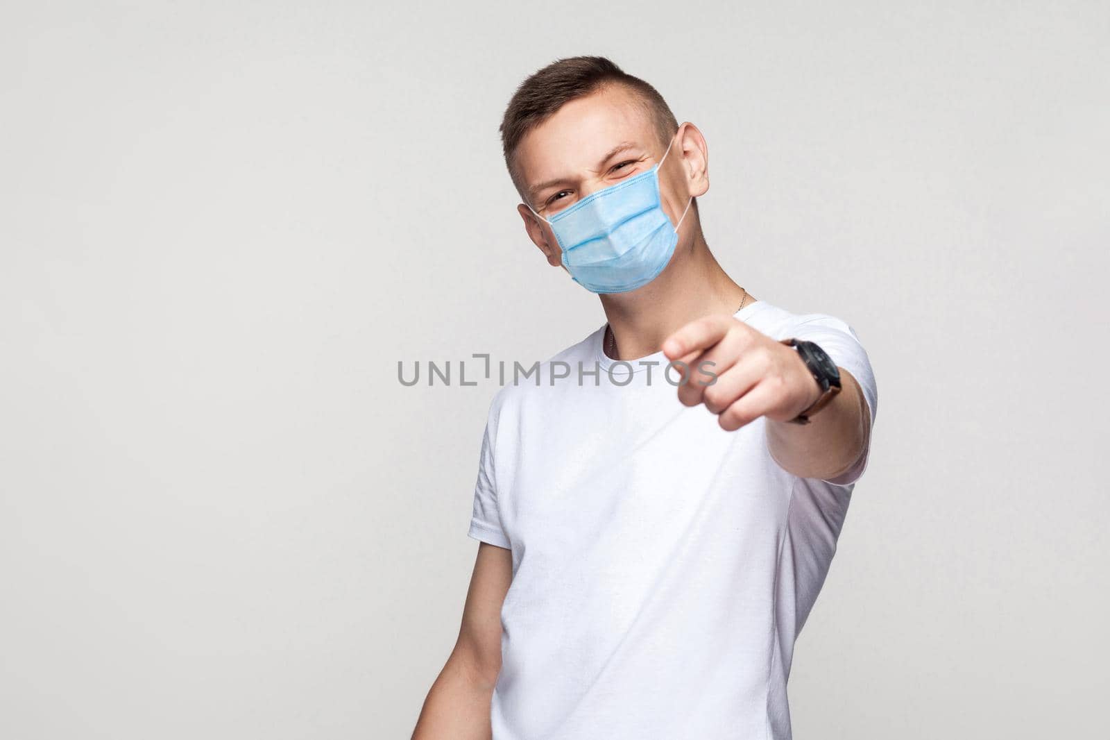 You are so funny. Portrait of young man in white shirt with surgical medical mask standing, looking and pointing at camera with smiley face. indoor studio shot, isolated on gray background.