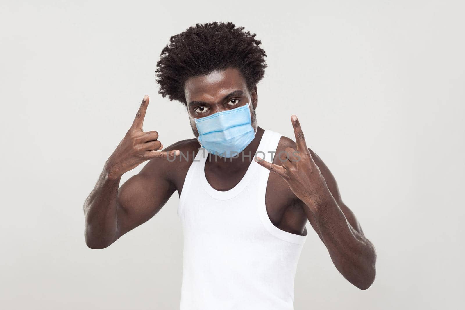 Portrait of young man wearing white shirt with surgical medical mask standing with rock horns gesture and looking at camera. indoor studio shot isolated on gray background.