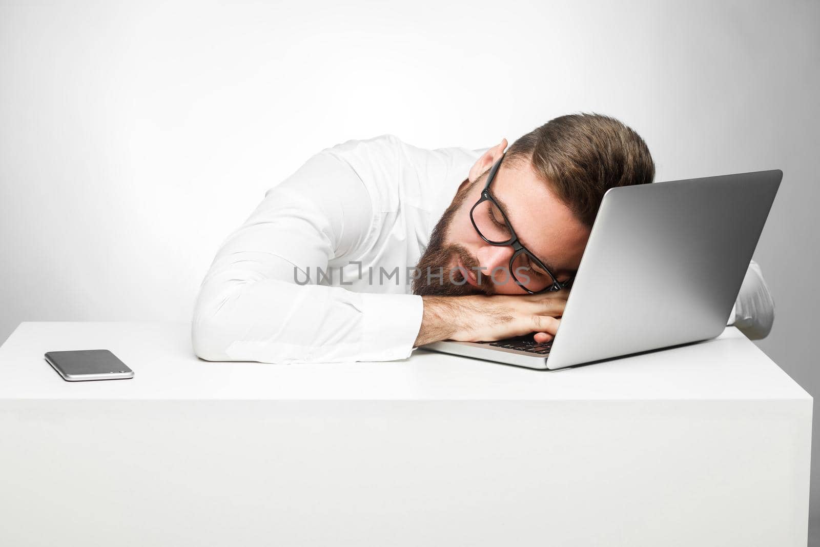 Sweet dreams in the work station. Portrait of sleepy tired freelancer in white shirt are sitting in office is snoozing at his work place near laptop. Indoor, studio shot, gray background, isolated