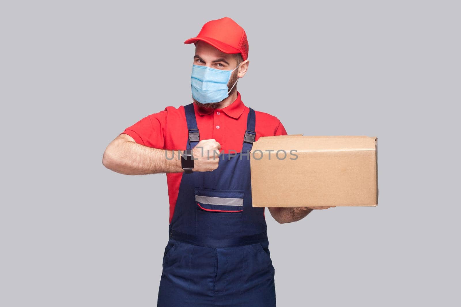 Delivery on quarantine. Ontime service! man with surgical medical mask in blue uniform and red t-shirt standing, holding delivery box and showing watch on grey background. Indoor shot, isolated,