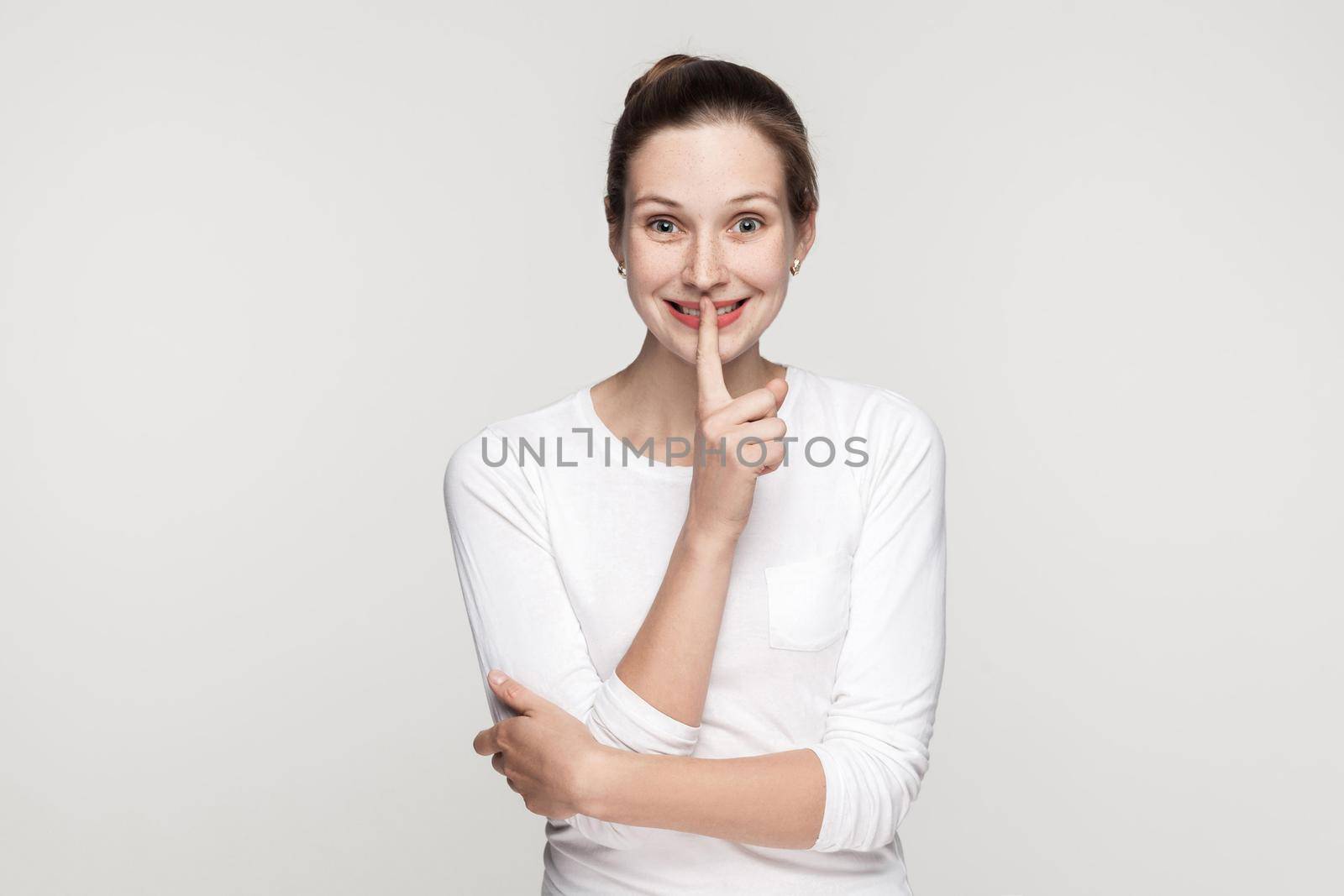 Shocked young woman, showing shh sing and looking with big eyes at camera and smiling. Studio shot, isolated on gray background