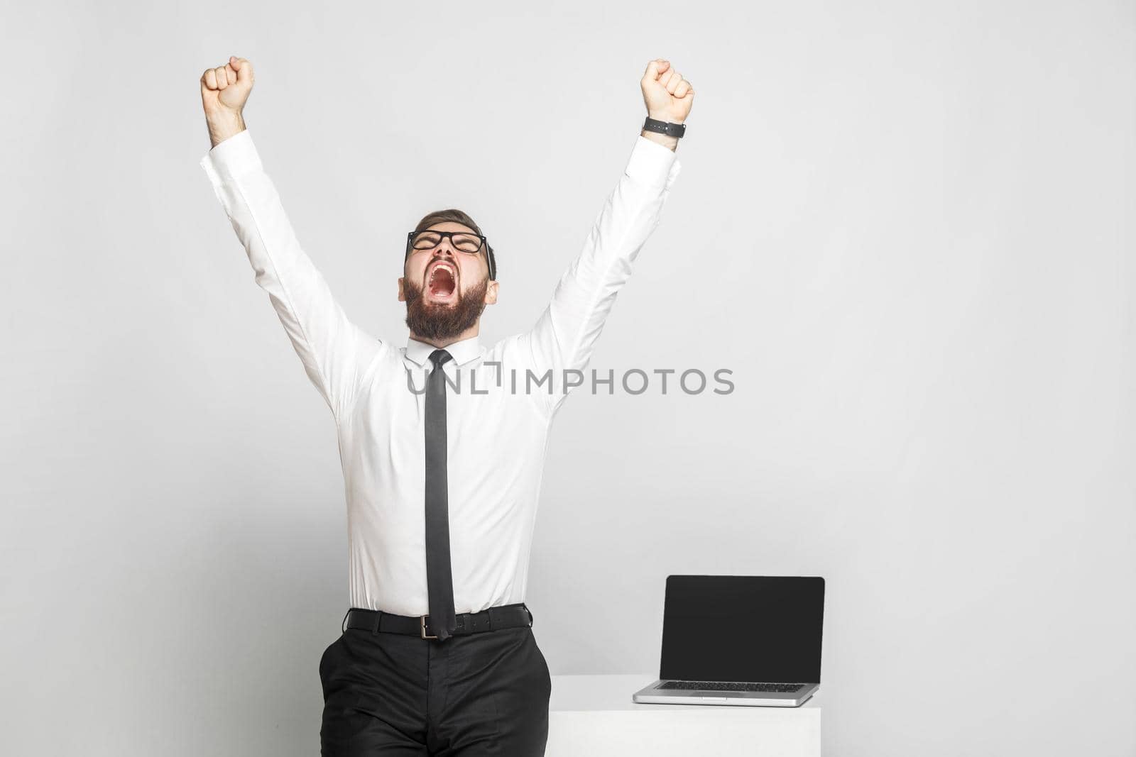 Yes! Happy screaming bearded young businessman in white shirt and black tie are standing near his working place are triumphing with open mouth and raised arms. Isolated, studio shot, gray background