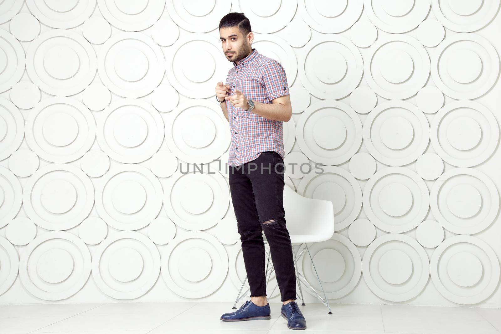 Young bearded fashion model in casual style is posing near white circle wall background. studio shot. looking at camera with pointing finger.