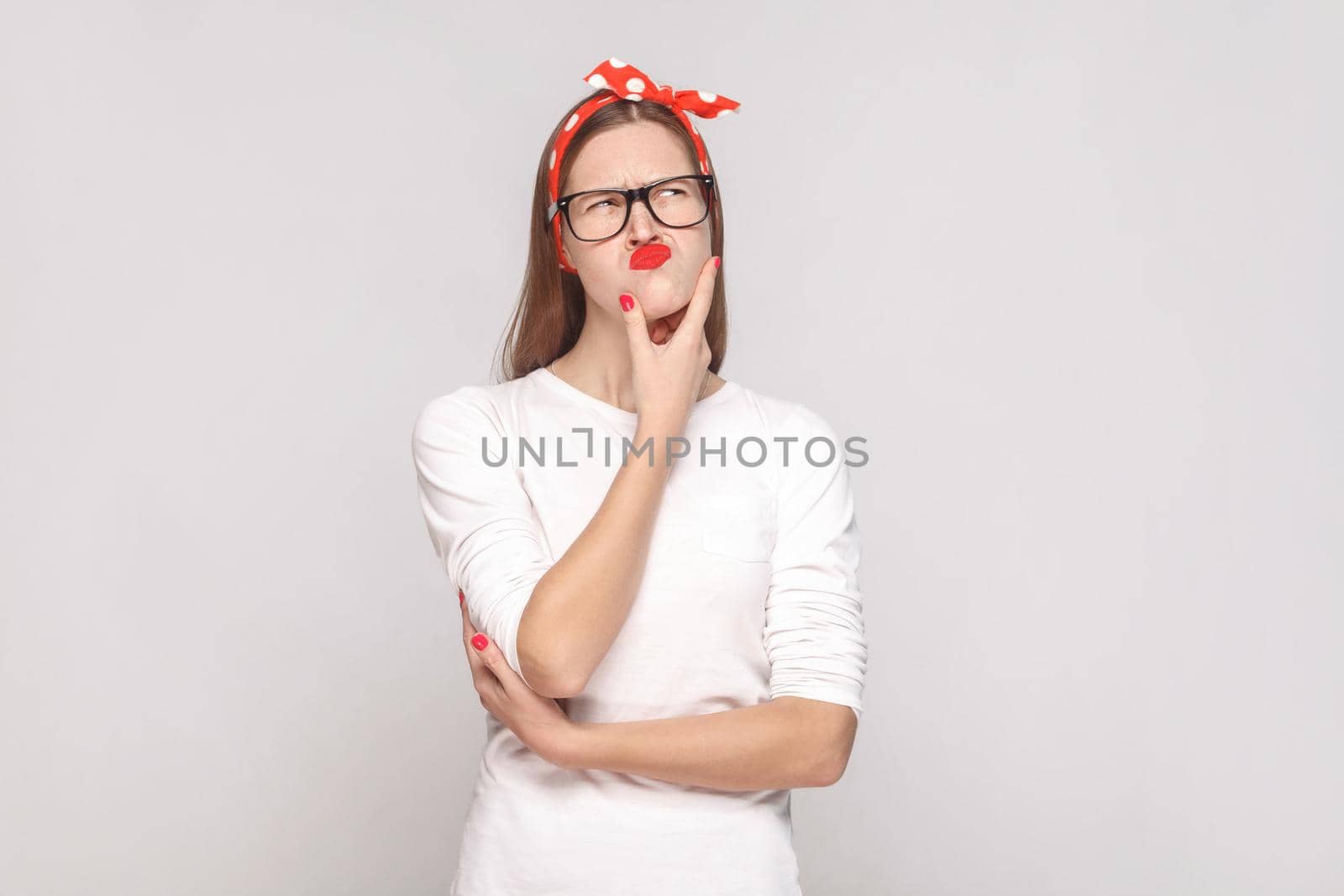 portrait of beautiful emotional young woman in white t-shirt with freckles, black glasses, red lips and head band touching her chin and thinking. indoor studio shot, isolated on light gray background.