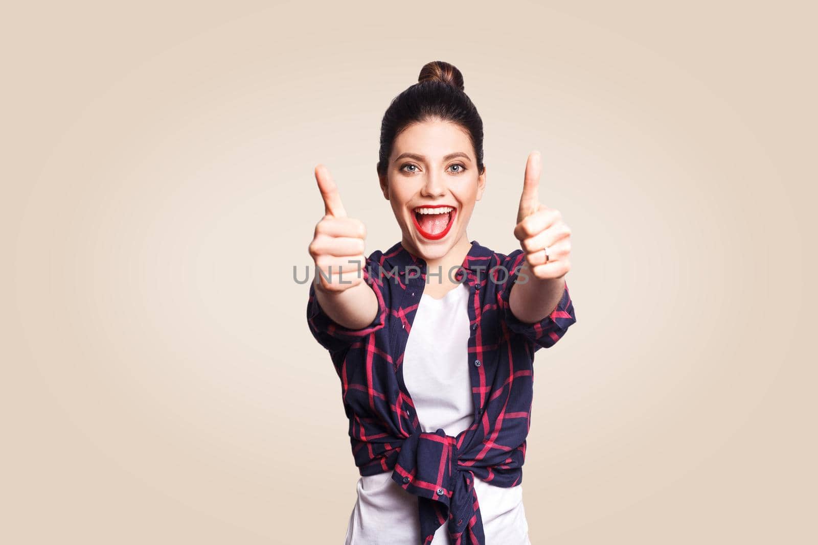 Young happy girl with casual style and bun hair thumbs up her finger, on beige blank wall with copy space looking at camera with toothy smile. focus on face.