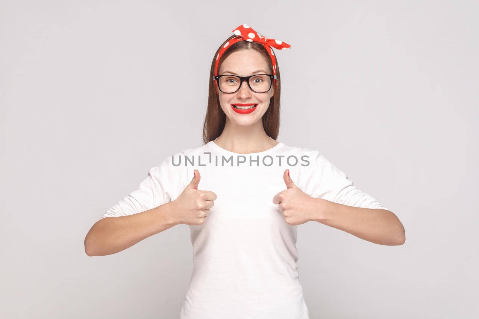 I am satisfied, thumbs up. portrait of beautiful emotional young woman in white t-shirt with freckles, black glasses, red lips and head band. indoor studio shot, isolated on light gray background.