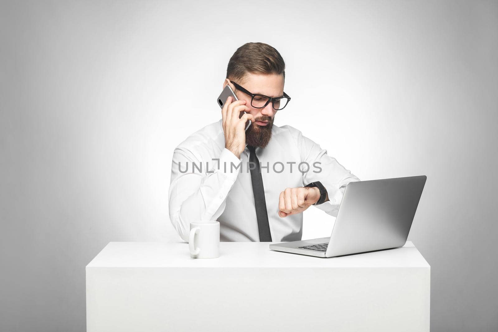 Thoughful bearded young worker in white shirt and black tie are sitting in office on desk and talking with partner on phone also checking time on his own hand watch, planning meeting. Gray background