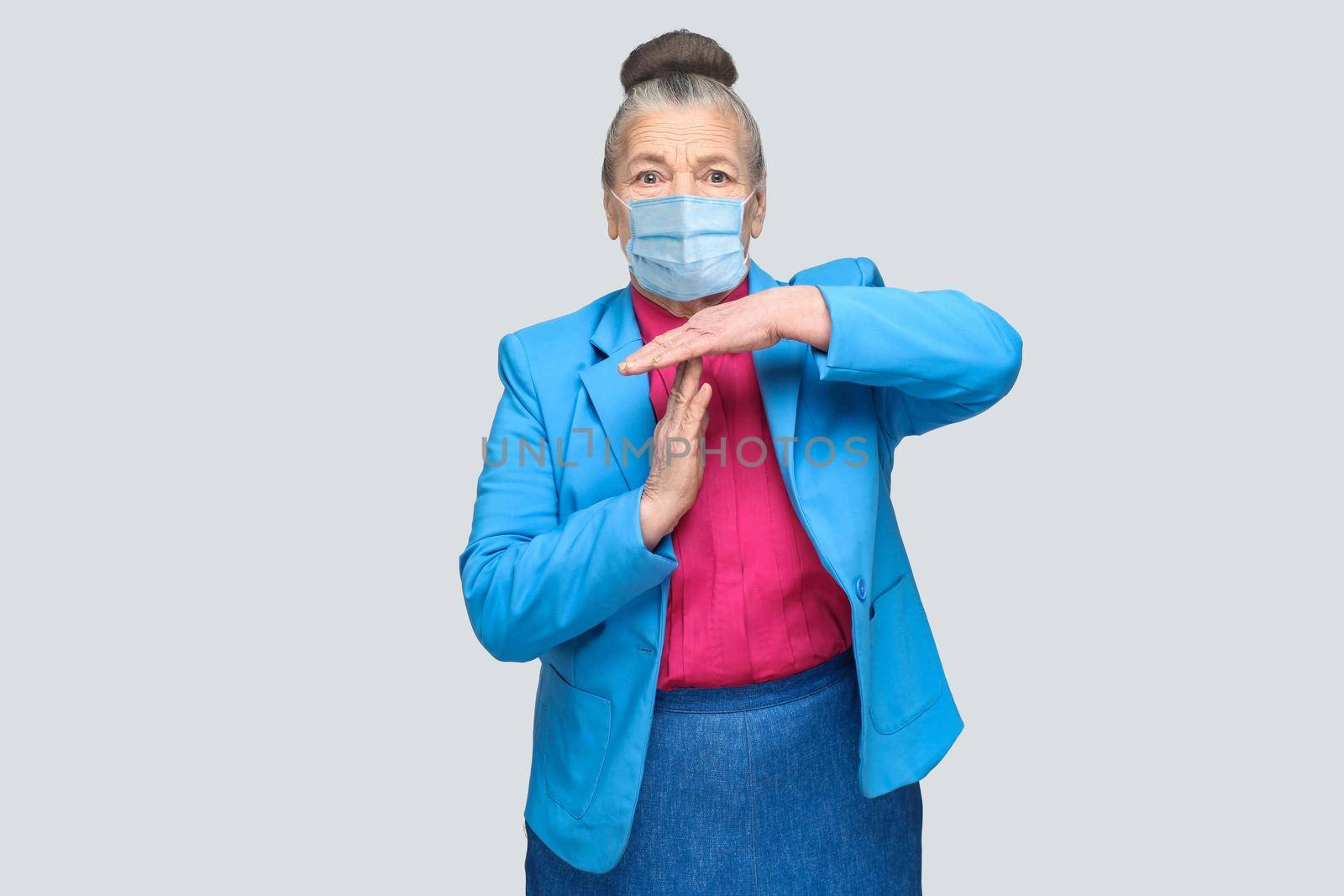 Aged woman with surgical medical mask showing time out sign. Grandmother with light blue suit and pink shirt standing with collected bun gray hair. indoor studio shot, isolated on gray background