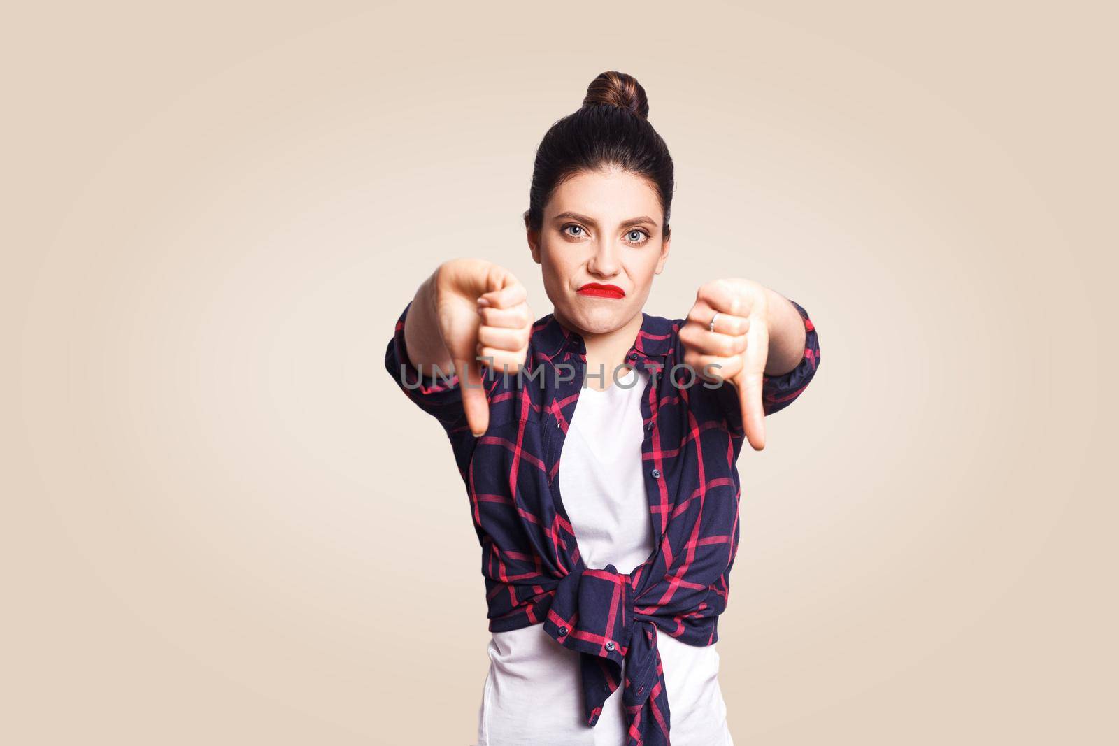 Dislike. Young unhappy upset girl with casual style and bun hair thumbs down her finger, on beige blank wall with copy space looking at camera with toothy smile. focus on face.