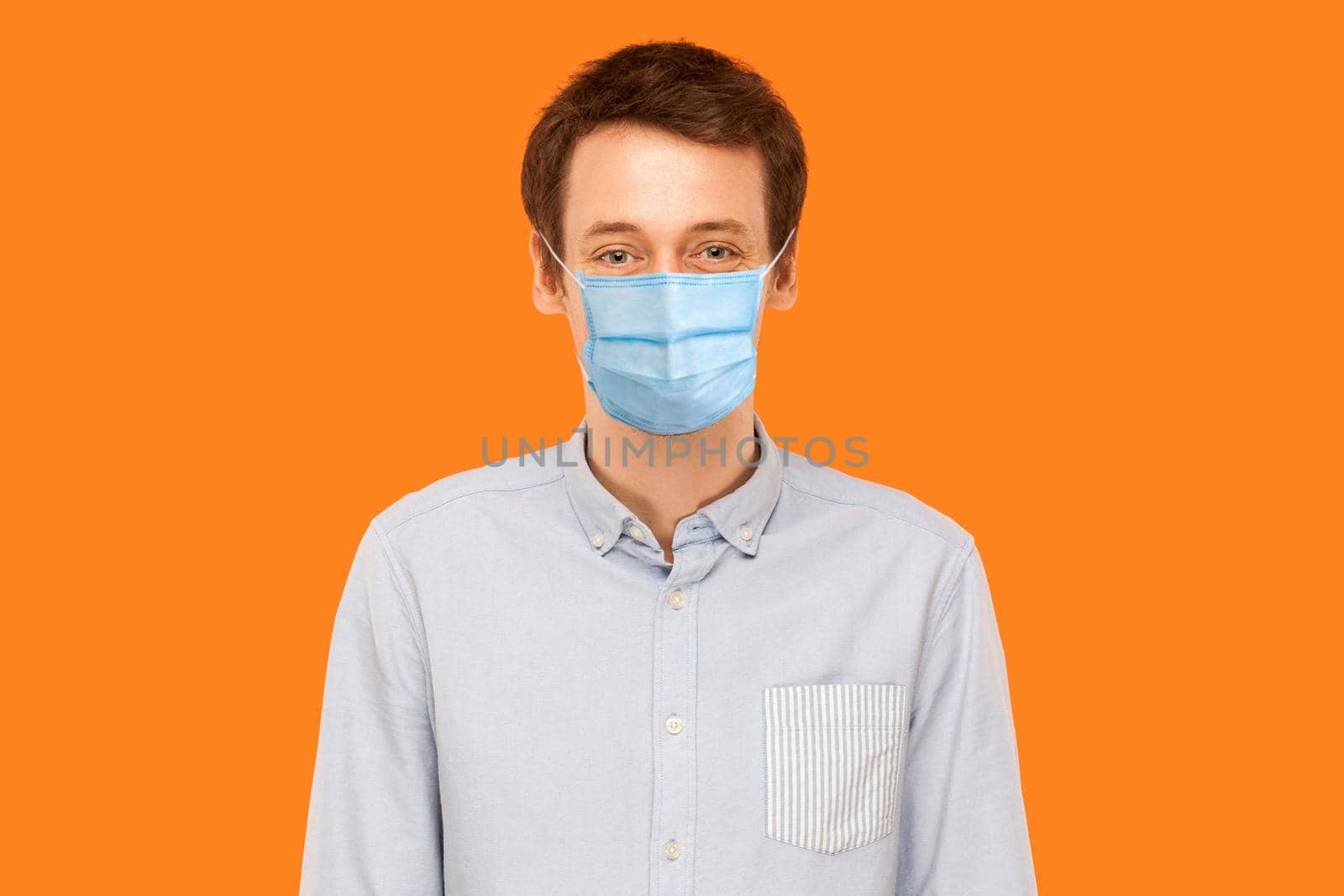 Portrait of young worker man with surgical medical mask standing and looking at camera smiling. health care and medicine concept. indoor studio shot isolated on orange background.