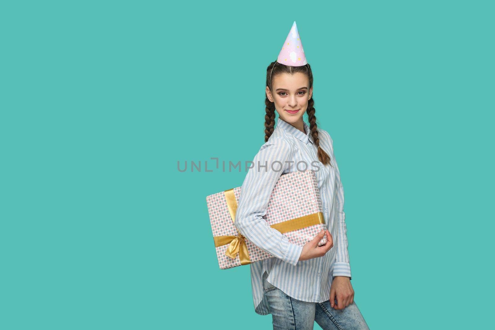 Happy funny girl in striped blue shirt standing with dotted gift box and birthday cap on her head, looking at camera with safisfied smiley face, Indoor studio shot, isolated on green background