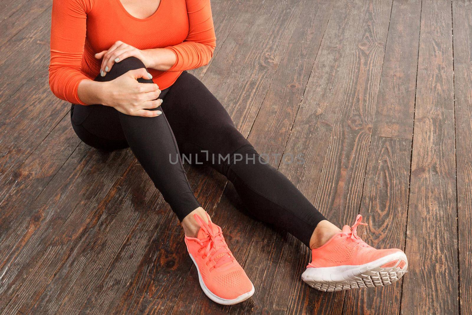 Fit woman in tight sportswear holding painful knee sitting on floor at home gym, suffering muscle strain, sprain ligaments or joint injury, health problems after sports training. indoor studio shot